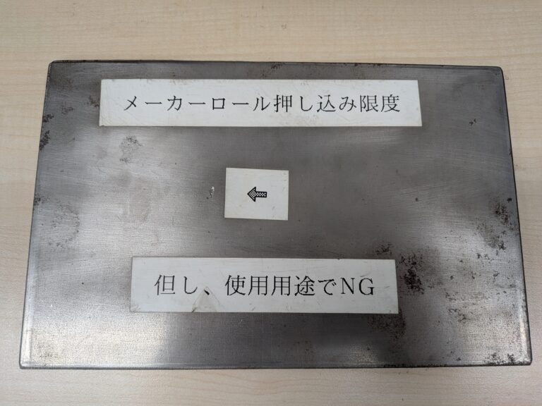 鋼板の表面写真。「メーカーロール押し込み限度」「但し、使用用途でNG」と書かれた札と矢印プレートが置かれているが、欠陥そのものは見分けにくい。