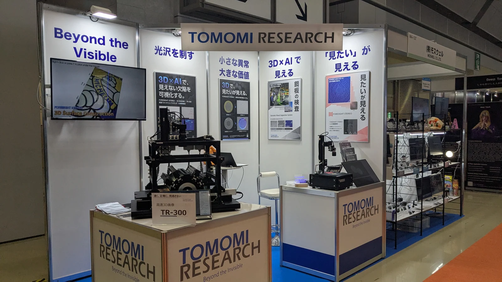 国際ロボット展 2025 の TOMOMI RESEARCH ブース。正面の壁に「Beyond the Visible」「光沢を制す」「小さな異常 大きな価値」「3D×AI で見える」「「見たい」が見える」と大きく掲出され、中央の台に多方向照明を組んだ高速 3D 画像装置 TR-300 が、右手の台に小型の観察装置が展示されている。