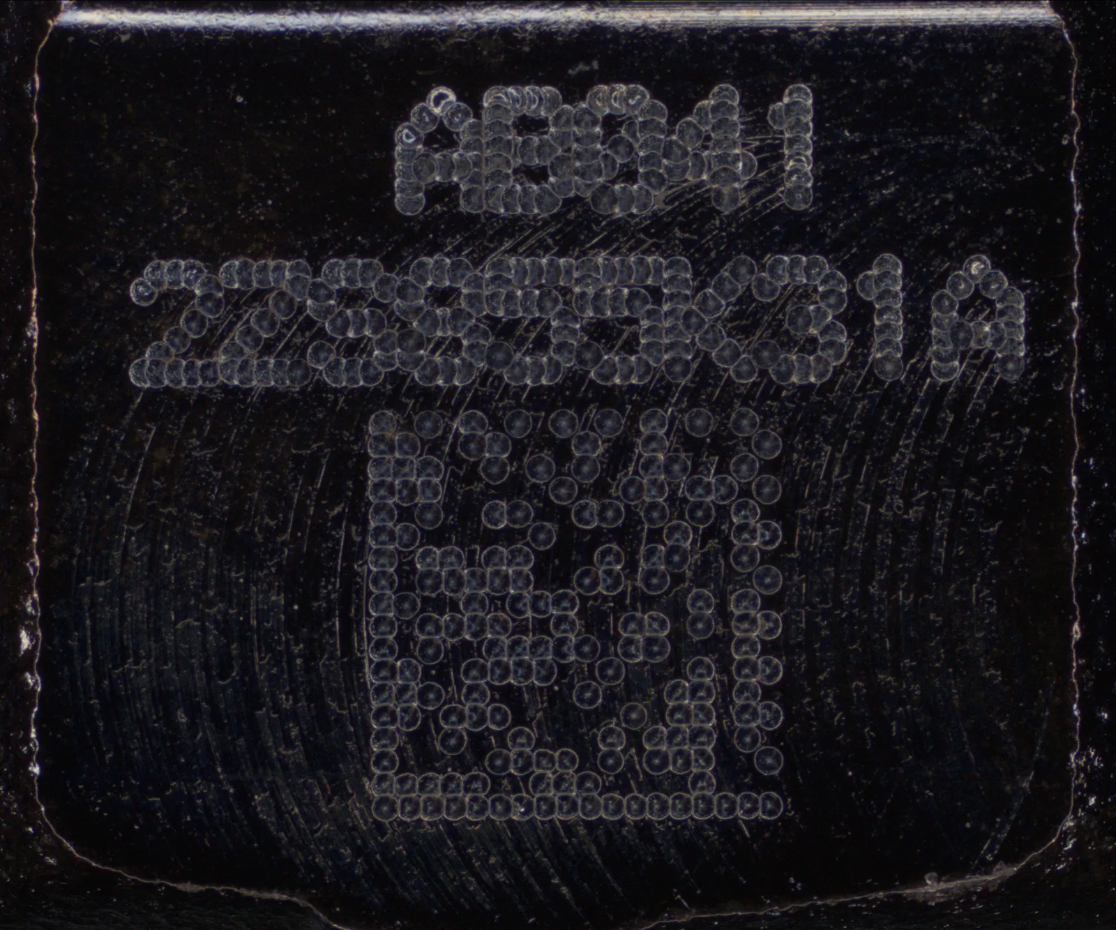 黒い部品に刻まれた識別記号と DataMatrix コードを通常のカメラで撮影した画像。面がほぼ黒く沈み、刻印はかろうじて分かる程度にしか読み取れない。