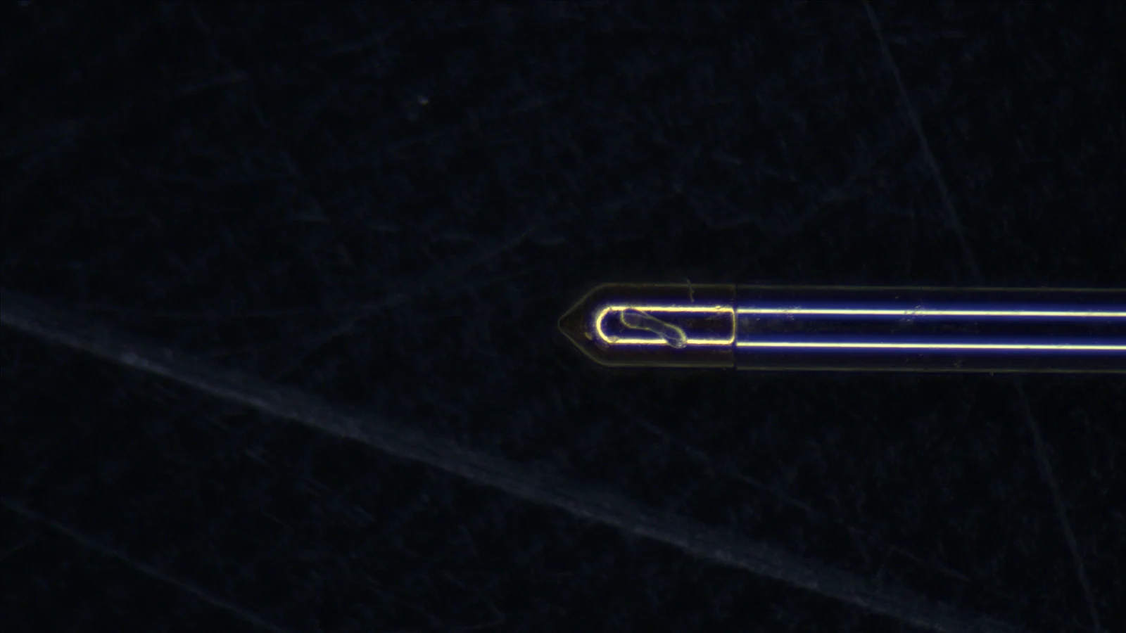 A machined metal face photographed with an ordinary camera. Almost no light returns, the whole frame sinks to black, and neither the machining grain nor the scratches can be read. Only the tip of a fine needle pointing at a scratch catches the light at the centre.