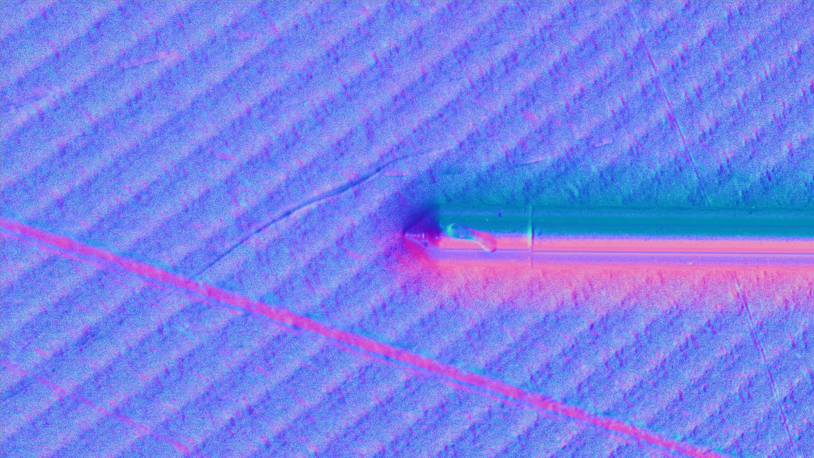 Surface Normal output of the same metal face. Machining striations run diagonally across the whole frame, and a single scratch crossing them, together with a short scratch indicated by a needle tip, separate clearly as a different surface direction.